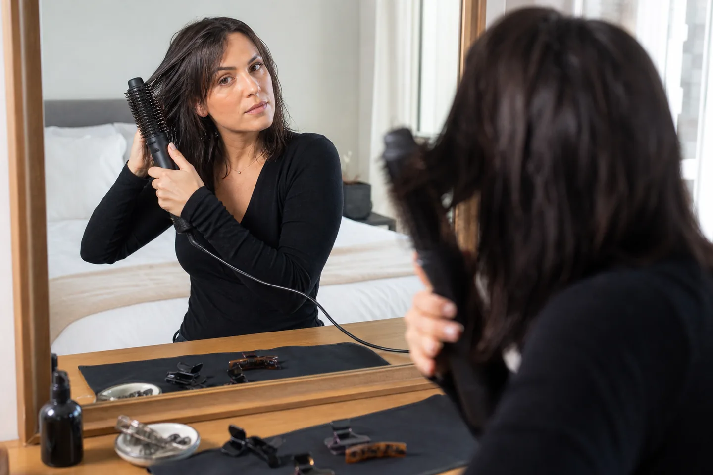 Woman using an unbranded blow-dryer brush on damp shoulder-length hair at a bedroom vanity