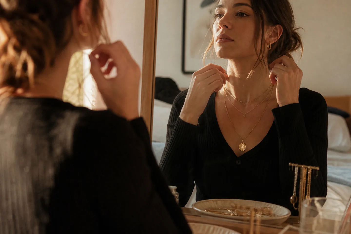 Woman layering simple gold necklaces and small hoop earrings at a warm bedroom mirror