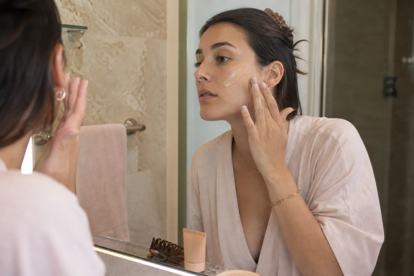 Woman applying sheer skin tint with her fingers at a bathroom mirror in natural morning light